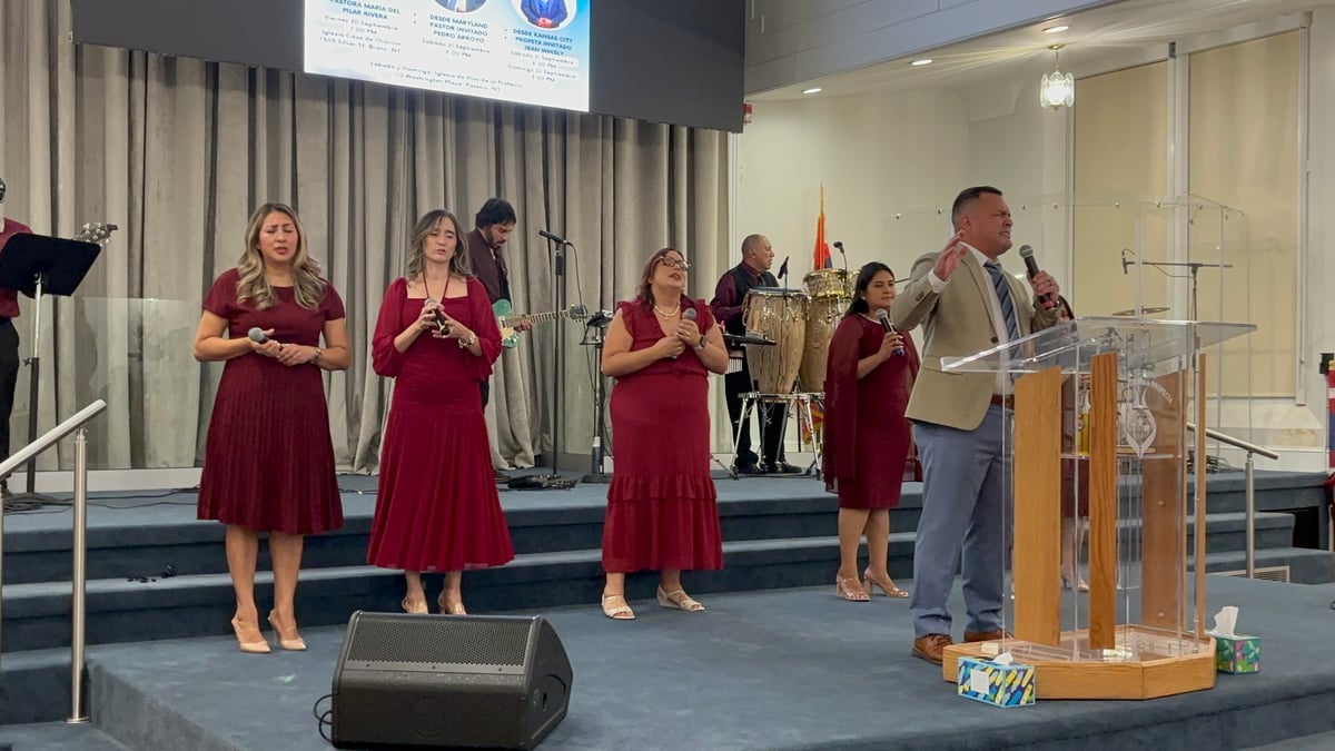 Pastor predicando desde el podio con grupo de mujeres en vestidos burgundy cantando en el escenario del Concilio Internacional Visión en Acción